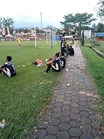 The video captures an outdoor scene at what appears to be a community event or sports tournament in Matesih, Indonesia. A soccer match is underway on a large green grass field, with multiple players in various jerseys visible. Two soccer goals are present on the field.

In the foreground, several spectators are gathered along a paved path next to the field. Some are sitting directly on the grass, while others are on the paved area. One person, seen from behind at the 0:02 mark, is wearing a dark jersey with the number "6" and possibly the text "TIMBUNG-SARI" or similar on the back, and is watching the game. Another group of people is sitting near a cardboard box. There are discarded plastic cups and other trash visible on the grass and path, suggesting a lively gathering.

In the background, beyond the soccer field, numerous white tents or stalls are set up, indicating a market, festival, or event area. Trees and some buildings are also visible, along with distant mountains or hills under an overcast sky. The lighting suggests it is late afternoon or early evening.