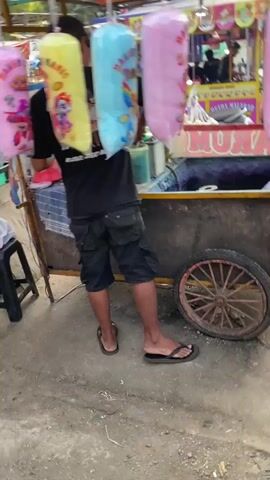 The video shows a street vendor preparing and selling cotton candy at an outdoor market. The vendor, a man wearing a black t-shirt and shorts, stands behind a cart piled high with colorful cotton candy. He is holding a large pink cotton candy bag. Several other large bags of cotton candy, yellow, blue, and pink, are hanging from the cart. The cart has a large red and white sign that reads "ARUM MANIS" which translates to "sweet fragrance" or "cotton candy." In the background, other vendors can be seen selling snacks and drinks. The scene appears to be in a bustling marketplace.