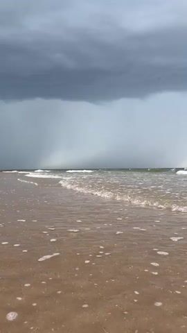 A wide shot from a beach looking out at the ocean. Dark, ominous storm clouds fill the upper half of the frame. Rain can be seen falling in sheets in the distance. Gentle waves lap against the sandy shore, leaving a trail of white foam. The scene is calm and serene, despite the approaching storm.