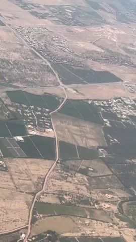 The video offers an aerial perspective of the Al Ouidane region in Morocco, viewed from an airplane window during what appears to be daytime. The scene showcases a vast, semi-arid landscape dominated by a patchwork of agricultural fields and scattered human settlements.

The ground is predominantly light brown and tan, characteristic of a dry environment, interspersed with numerous green plots indicating active cultivation. Many fields feature discernible rows, suggesting orchards, vineyards, or other crops. Other fields appear fallow or dry. A network of dirt roads and possibly some paved routes crisscrosses the terrain, connecting various clusters of buildings and individual houses that form small villages or hamlets. The settlements are spread out across the landscape, often adjacent to or within the cultivated areas.

The sky is clear and blue, with some atmospheric haze visible in the distance. The overall view provides a serene and expansive look at human land use and habitation patterns within an arid region. No people or visible text are present in the footage.