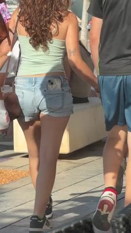 The video shows a group of people walking outdoors. In the foreground, a person wearing blue shorts and a green t-shirt walks on the left. To their right, a person in denim shorts and a green tube top walks, holding a doll and a phone in their back pocket. On the far right, a person in black shorts and a dark t-shirt walks, holding hands with the person in the denim shorts. The environment appears to be a paved walkway with some greenery and buildings in the background. The lighting suggests it is daytime.