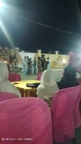 The video shows a group of people dressed in traditional attire dancing in what appears to be an outdoor event space at night. There are several people sitting in pink chairs watching the dancers. The dancers are moving and interacting with each other, while the seated individuals are observing the performance. The environment is lit with artificial light, and there are draped fabrics and decorations in the background. A table with a gold tablecloth is visible in the foreground.