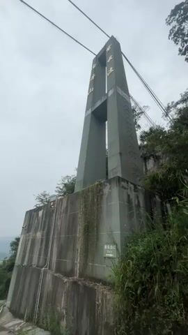 The video opens with a low-angle shot of a tall, green pylon supporting thick cables, likely part of a bridge or cable car system. The pylon is built on a large, textured concrete base overgrown with moss and vegetation. Golden Chinese characters are visible near the top of the pylon. To the right, dense green foliage climbs the side of a rocky incline.

The camera then pans left, revealing a road leading towards the structure. The sky is overcast.

As the video progresses, the camera moves onto a pathway or viewing platform with a railing. Several people are present, some wearing brightly colored vests. A sign with Chinese text and images is mounted on the railing. In the background, a valley with trees and distant mountains can be seen under the cloudy sky. The cables continue to stretch overhead, disappearing into the distance. The video ends on this panoramic view of the bridge structure and surrounding landscape, with people milling about on the platform.