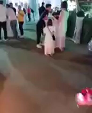 This media file captures a lively outdoor event, likely a celebration or gathering, taking place at dusk in Kırıkkale, Turkey. The scene is illuminated by artificial lighting, creating a festive atmosphere.

The primary subjects in focus are several people, including adults and a young child dressed in a white dress. They appear to be interacting around a table adorned with a white tablecloth, suggesting a reception or a buffet area. One adult, dressed in black, is leaning towards another adult who is wearing a light-colored dress. The child in white stands between them, observing the interaction. In the background, more individuals are visible, mingling and enjoying the event.

The setting appears to be a well-decorated outdoor venue, possibly a garden or a courtyard. There are white pillars and an arched structure visible, suggesting a formal or semi-formal setup. Green lights are casting an ambient glow on some of the surroundings, enhancing the celebratory mood. A neon sign with green text is partially visible in the background, though the text itself is not clearly legible.

The overall mood seems to be one of joy and social engagement, characteristic of a wedding reception, birthday party, or a similar festive occasion. The time of day, indicated by the dim natural light and prominent artificial lighting, suggests the event is taking place in the evening.