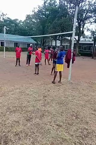 A group of around ten people is playing volleyball on a dirt court. Some of the players wear red uniforms, while others wear blue and yellow or maroon outfits. The volleyball net is strung between two white poles. Behind the court are green trees and a few single-story buildings with light-colored roofs. The ground surrounding the court is covered in dry grass. The players are hitting the volleyball back and forth over the net, some jumping to spike the ball.