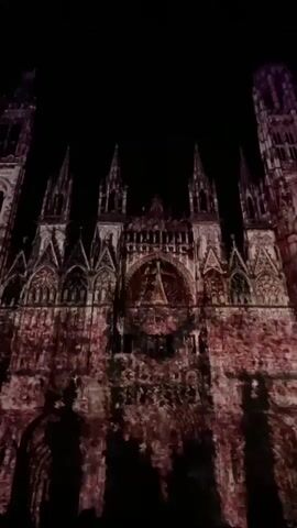A low-angle shot presents the ornate, dark facade of a large Gothic-style cathedral at night. The cathedral is illuminated by a light projection displaying a dynamic, textured pattern in shades of reddish-brown and dark tones across its entire surface, giving the appearance of aged or painted stone. This projected light subtly shimmers and shifts. The facade features multiple spires, arches, windows, and intricate architectural details. Orchestral music plays throughout the video.