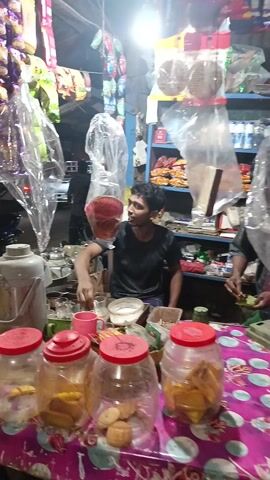 The video shows a bustling street scene at what appears to be a small shop. The camera focuses on a display of glass jars filled with various food items, each with a red lid. Behind the jars are a gray thermos and other containers. Racks of bagged goods, possibly snacks or candies, hang in the background. The shop seems cluttered with various items. The camera then moves, showing a narrow passageway with people walking through. One person is wearing a yellow shirt. The passage leads further into the shop or another part of the market, with more activity visible in the distance. The environment seems lively and crowded.