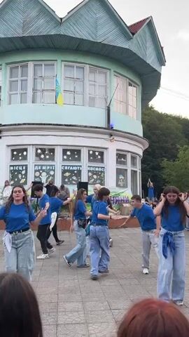 A group of young people in blue t-shirts and jeans are dancing outside a building with a light green and white facade. The building has several windows, some decorated with posters depicting stars and planets. A Ukrainian flag is visible hanging from the building. In the background, a few other people are standing and watching. Some of the dancers are in the foreground, and others are further back near a stage area where a person appears to be playing an instrument. The ground is paved with square tiles. The sky is overcast.