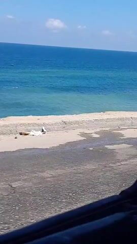 The video shows a man sitting on a wall by the ocean on a sunny day. The camera moves forward and the man leaves the frame. The view is then only of the ocean, the wall, and the road in front of it. The sky is blue with scattered white clouds.