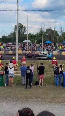 The video shows a demolition derby event. In the foreground, spectators are gathered behind a fence, watching the event. A few individuals in red shirts and yellow helmets, possibly event staff or safety personnel, are positioned near the track. In the background, a demolition derby track is visible with several wrecked cars. Two cars are currently on the track, one red and one blue, both appearing heavily damaged. Spectators are seated in bleachers surrounding the track. The environment appears to be an outdoor event with a cloudy sky.