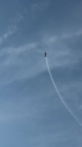 A small red and white airplane performs an aerial maneuver, leaving a white contrail in a clear blue sky with wispy clouds. The airplane is seen at different points in its curved trajectory.