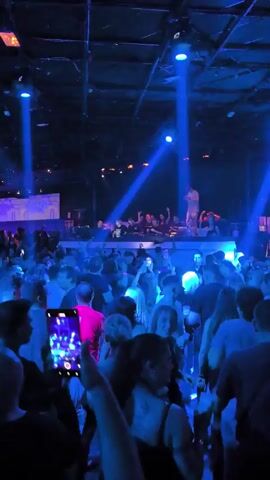 The video shows a crowded nightclub with people dancing and recording on their phones. A group of people, possibly DJs or performers, stand on a raised platform at the front. The club is dark, illuminated by flashing blue and purple lights. The atmosphere appears energetic and festive.