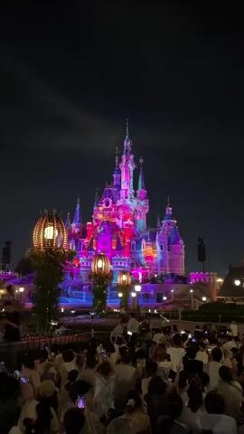 The video shows a nighttime view of a brightly lit castle, which is illuminated with colorful lights in shades of pink, purple, and blue. The castle has multiple spires and towers, and is surrounded by trees and lampposts. In front of the castle, a large crowd of people is gathered, most of whom are holding up their phones, presumably to record or photograph the spectacle. The sky is dark and appears to be cloudy. The overall scene suggests a fireworks show or a light projection event at a theme park, likely Disneyland, given the iconic castle structure.