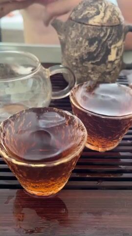 The video shows a close-up of a tea ceremony. In the foreground, two small, textured, amber-colored tea cups filled with dark amber liquid sit on a dark wooden tray with horizontal slats. Behind the cups, to the left, is a clear glass teapot with a handle. To the right of the glass teapot and slightly behind it is a small, stone-colored teapot with a dark lid and handle. A person's hands are visible in the upper left background, holding something out of frame. The lighting is natural and bright, suggesting an outdoor or well-lit indoor setting.