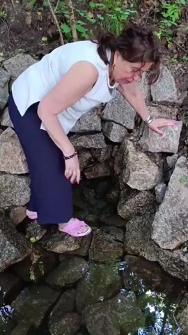 A woman with dark hair is bending over a small stream of water, which is flowing between large rocks. She is wearing a white shirt and navy pants, with pink sandals on her feet. Her right hand is reaching out towards the rocks, and her left hand is resting on a rock. The stream appears to be in a natural setting, with trees and foliage visible in the background. The water is clear, and the rocks are covered in moss and lichen. The scene suggests a warm day, possibly in the summer or early autumn, given the greenery. There is a wooden plank in the foreground, partially submerged in the water. The woman's posture and the way she is interacting with the water suggest curiosity or perhaps she is examining something in the stream. No text is visible in the image. The location context is Sdt Shcherbakovka, Russia.