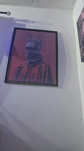 The video shows a corner of a room with two framed pictures hanging on the walls. On the left wall is a framed picture depicting a person wearing a crown and a dark covering over their face, dressed in a red patterned garment, against a red background. On the right wall is a larger, rectangular framed picture in gray tones. It depicts a scene with a figure kneeling and reaching upward, a figure lying prone, and other objects rendered in a stylized manner. The walls are white, and a section of a ceiling with a recessed light is visible above. The camera pans up to provide a wider view of the picture on the right wall.