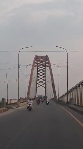 A wide shot shows a road leading onto a bridge. The bridge has a large, red, arched support structure with cables that extend down to the road. There are several motorcycles on the road, traveling in the same direction as the camera. The sky is overcast.