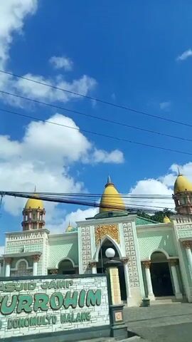 The video shows a green and white building with yellow domes, possibly a mosque, under a bright blue sky with white clouds. The building has ornate decorations with gold accents, and a sign that is partially visible on the left. Several electrical wires are strung across the top of the frame. The camera appears to be moving, possibly from inside a vehicle, as the perspective shifts slightly. Trees and other structures are visible in the background.
