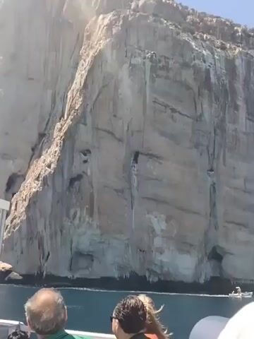 A video shows a boat on the water near a large cliff face. On the boat, several people are visible from the shoulders up, looking towards the cliff. One person in the foreground is wearing a hat. In the distance, another small boat is moving along the water. The cliff is tall and appears to be made of rock, with visible crevices and stains. The sky is clear and blue.