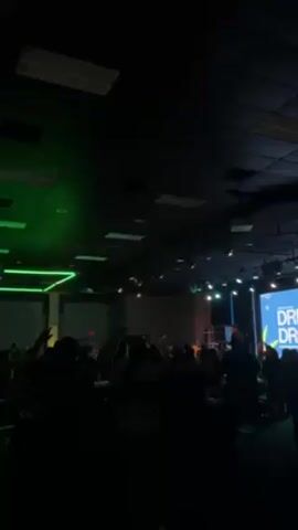 The video shows a gathering of people in a large room, possibly an auditorium or event hall, with a stage in the background. The room has a dark ceiling with rectangular light fixtures, some emitting a green light. People are facing the stage, many with their arms raised, suggesting they are participating in an event. The stage has a large screen displaying text and a performance area where figures can be seen, possibly musicians or speakers. Some tables are visible in the foreground. The overall atmosphere is dark with focused lighting on the stage, indicating a performance or presentation.
