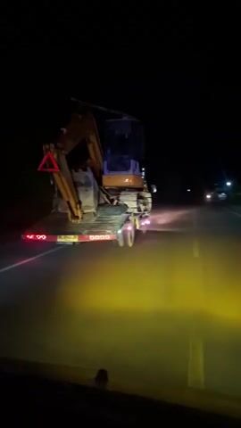 The video is filmed from the perspective of a vehicle at night, following another vehicle that is transporting a yellow and blue excavator on a flatbed trailer. The trailer has red lights on the back and purple lights on the underside. The road is dark with yellow lane markings illuminated by the headlights of both vehicles. Trees are visible on the sides of the road. The following vehicle's headlights are casting a yellow glow on the road ahead.