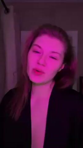 A medium shot of a fair-skinned woman with reddish hair, illuminated by pink lighting, facing the camera. She is wearing a black top. Her facial expression is neutral, and she is speaking or singing. The background is blurred but appears to be an indoor space.