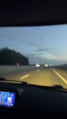The video shows the interior of a car driving on a highway at dusk or night. The camera is positioned behind the steering wheel, looking out through the windshield. The dashboard is visible in the foreground, with a navigation screen displaying a map and a car's interior lights on. A person's hand is visible, touching a car stereo. The road ahead is illuminated by the car's headlights and the headlights of other vehicles in the distance. Trees and vegetation line the sides of the highway. The sky is a deep blue, indicating it is either early evening or late night.