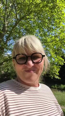 A fair-skinned woman with short blonde hair is centered in the frame, wearing black-rimmed glasses and a brown and white striped shirt.  She faces the camera with a slight smile. Behind her is a park-like setting, featuring green grass, mature trees with bright green leaves, and a clear blue sky. A small, newly planted tree is visible with a wooden support structure around it. The video appears to be taken in bright daylight.