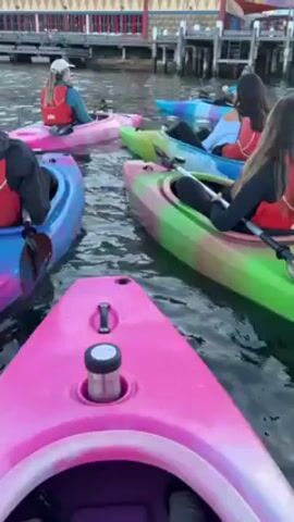 A group of at least six people is kayaking in what appears to be a harbor or bay.  Each person is in their own brightly colored kayak, mostly pink, green, and blue.  All are wearing red life jackets.  They are clustered together in the water, close to a dock or pier structure.  There is a tall, colorful structure in the background, possibly a roller coaster or other amusement park ride.  The water is slightly choppy.  The camera is mounted on one of the kayaks, providing a first-person view.  The overall impression is that they are on a recreational kayaking trip.