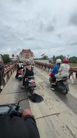 The video shows a view from the perspective of a motorcycle rider on a bridge. Several other motorcycles, some carrying multiple passengers, are also on the bridge, creating a line of traffic. In the distance, a large truck is visible. Indonesian flags are flying on poles along the side of the bridge. The sky is cloudy. The bridge appears to be made of metal and wood. The road surface is wet and appears to be made of metal plates in some sections. The rider's hand is visible on the handlebars of the motorcycle in the foreground.