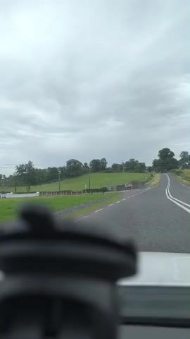 This is a point-of-view shot from inside a vehicle, looking forward through the windshield on a cloudy day. The road ahead is paved and appears to be a rural route, curving gently to the right. The landscape on either side of the road is lush green, with fields and scattered trees. In the distance, a large, mature tree stands prominently on the right side of the road.

Several signs are visible along the left side of the road. One sign, predominantly green, has white text and appears to be informative, possibly directional. Further along, a brown sign with white lettering is also visible, seemingly indicating a local destination or attraction. There are also utility poles and wires running alongside the road.

The foreground of the image is dominated by a blurry, dark object, likely a piece of equipment mounted on the dashboard, such as a GPS device or a dashcam. This object obscures the immediate interior of the vehicle.

The weather is overcast, with a thick layer of grey clouds obscuring the sun, suggesting it could be morning or afternoon. The overall mood conveyed is one of a quiet, potentially uneventful drive through a verdant rural setting. The location cues, such as the style of road, fencing, and the green landscape, are consistent with a rural Irish countryside. No people are visible in the frame.