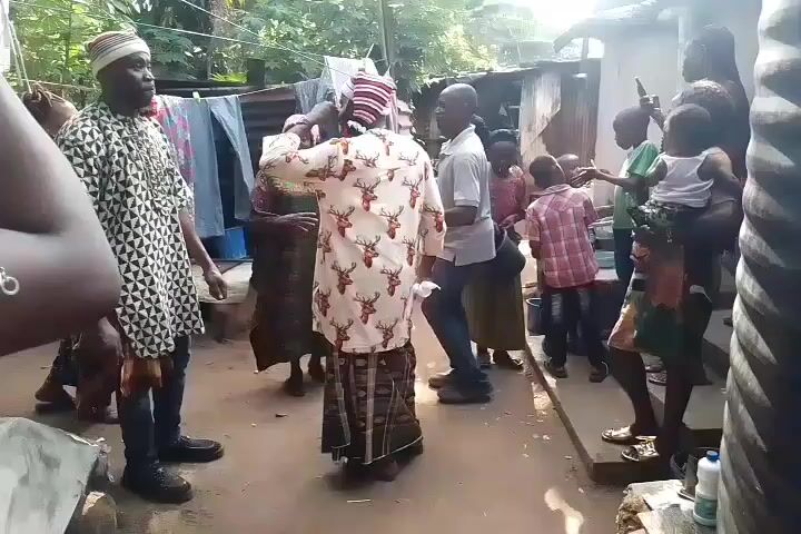 The video shows a group of people gathered outside, with music playing in the background. Some people are dancing, including a person in a deer-print shirt and a striped head wrap. There are children among the crowd. A man in a patterned shirt and cap stands to the side, watching the dancers. Clothes are hanging on a clothesline in the background.  The setting appears to be a residential area in a developing country, given the corrugated metal structures and simple surroundings.