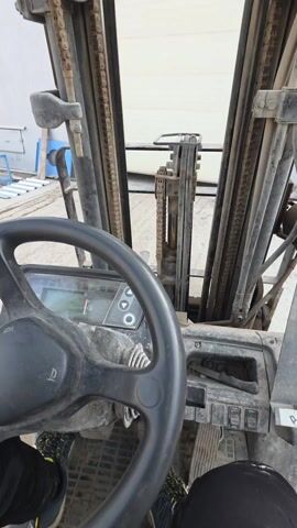 The video shows the interior of a forklift, focusing on the steering wheel and mast assembly. The steering wheel is black and shows signs of wear, while the mast is made of metal with visible chains and hydraulic lines. Through the forklift's front, there's a view of a building exterior with a large door, and metal structures can be seen in the background. The forklift appears to be dirty and used. A person's legs are visible at the bottom of the frame, indicating the operator's perspective.