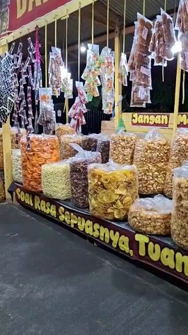A low-angle shot shows a brightly lit snack stall at night with numerous large, clear plastic bags filled with various chips, crackers, and snacks displayed on a counter. Above, many smaller packaged snacks hang from the ceiling. A young man with dark hair, wearing a black hooded jacket and dark pants, stands behind the counter, focused on a string of small, yellow mesh bags, each containing colorful, round candies, which are also hanging from the ceiling. He appears to be arranging or untangling them. A yellow sign with partially visible Indonesian text "Soal Rasa Sepuasnya, Tuankuu" and "Jangan Malu', OK" runs along the bottom of the counter and on the back wall respectively.