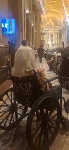 The video shows a church sanctuary from the perspective of someone seated in a wheelchair. The church is well-lit and ornately decorated with a golden altar. There is a large crowd of people seated in rows of pews, some standing, and others walking through the aisles. In the foreground is an elderly woman with white hair seated in a wheelchair. The wheelchair has "ALICIA SALUD 5" printed on the back. People are milling around. The camera pans slightly to give a better view of the church interior. A small TV monitor is visible in the top left corner of the frame.