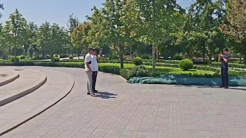The video shows a park with a large equestrian statue of Amir Temur. Two men in white shirts and dark pants are standing near the statue. One man is looking at the statue, and the other is looking away. There are trees and bushes in the background, and the sky is clear and blue. The ground is paved with stone tiles. There are also some signs and red ropes around the statue. Some green netting is covering some of the bushes.