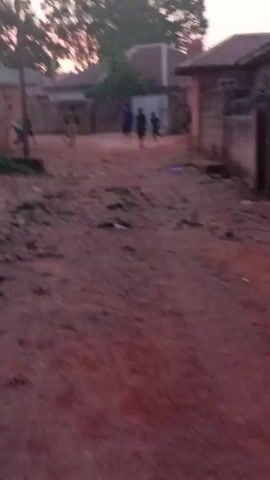 The video shows a dirt road in what appears to be a village or rural area, with several people, mostly children, walking or running down the road. The road is red dirt, and there is some debris scattered on it.  Houses line the sides of the road, some made of brick or concrete. The sky is visible, showing a sunset or twilight. The camera appears to be handheld, and there's some shakiness in the video.
