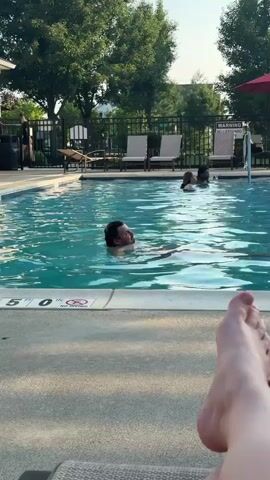 The video shows a man swimming in a pool. He is wearing swim trunks and his head and shoulders are visible above the water. The man has dark hair and a beard. He is looking towards the camera. The pool is surrounded by lounge chairs and trees. There are other people in the pool in the background. The video is shot from a first-person perspective, suggesting the camera is being held by someone relaxing by the pool.