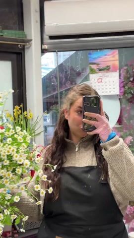 A woman stands in a flower shop, holding a large bunch of white and yellow daisies in front of her. She is taking a photo of herself with the flowers using her smartphone. She wears a brown sweater, a black apron, and pink gloves. The background shows the interior of the flower shop, including a window with a calendar, other flowers, and shop fixtures.
