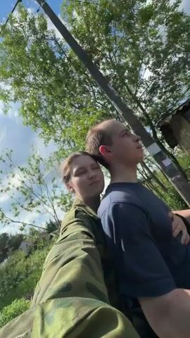 A fair-skinned young woman wearing a camouflage jacket and a fair-skinned young man wearing a navy blue T-shirt are riding a motorcycle on a sunny day. The camera is held by the woman, who is in the foreground, with the man in front of her. The woman is holding on to the man and resting her head on his back. The man is looking forward. The background shows trees, a fence, and a blue sky with white clouds.