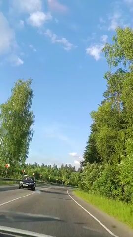 This video shows a first-person perspective of driving on a paved road that curves to the right. The road is bordered by lush green trees and foliage on both sides. The sky is clear blue with scattered white clouds. On the left side of the road, there are several red and white striped traffic markers. The trees on the left appear to be a mix of deciduous and coniferous species, while the trees on the right are predominantly coniferous, with tall pines. The sunlight suggests it is daytime. The overall impression is a scenic drive through a wooded area.