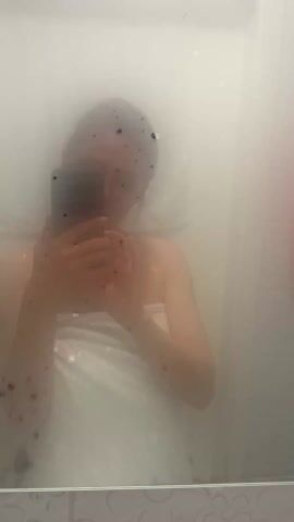 The video shows a person taking a selfie in a steamed-up bathroom mirror. The person is partially visible, wrapped in a white towel. A phone is held in their hands, obscuring some of their face. The mirror is speckled with water droplets. A pink towel hangs on a hook in the background to the right. The overall lighting is soft and diffuse due to the steam.