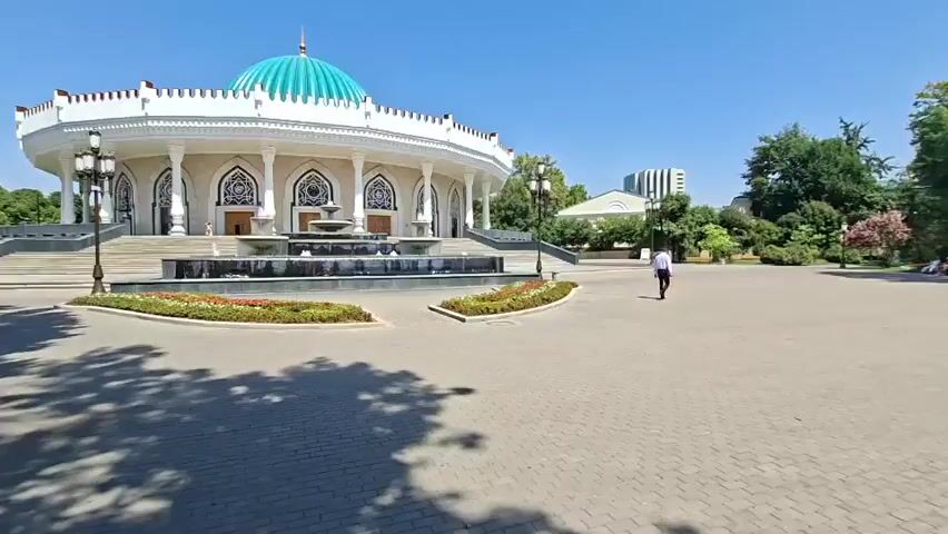 A wide shot of a building with a turquoise dome, possibly a palace or museum, on a clear sunny day. The building is ornate and white with columns and intricate carvings. In front of the building is a tiered fountain with water jets. The area around the fountain is paved with decorative flower beds. A paved walkway leads from the foreground toward the building. Several trees cast shadows on the walkway. A man in a white shirt and dark pants walks away from the camera towards the building. In the background, other buildings and trees are visible. The overall atmosphere is serene and grand.
