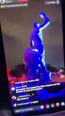 The video displays a phone screen with a live stream playing in a vertical format. The live stream captures an outdoor night scene with bright illuminations. In the foreground, there are chat messages overlaid on the video. The first chat message, "C'est quoi de l'autre côté," is visible at the bottom of the screen. Above this, a user named "Nesbeal089" has commented, "Lindsay c'est quand les franc maçons ont fait tomber la royauté française." Another user, "Chrâamel la," has written, "Gey fenm la." A user named "Mme Konaté" has joined the stream. The live stream itself shows a large, illuminated structure, possibly a statue, with blue and white lights. Further in the background, the Eiffel Tower is visible, also lit up, with what appears to be a light show or fireworks in the sky around it, creating swirling patterns of light. A crowd of people is gathered at the base of the Eiffel Tower, illuminated by the lights. The overall environment appears to be a public space at night, likely a celebration or event. The top of the phone screen shows a username "SantosSimpk" with "5.7K" and a "Suivre" button, indicating it is a social media live stream. There are also icons for likes and comments.