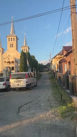 The video shows a wide, street-level view of a church with three spires. A white minivan is parked in the foreground, facing away from the camera. To the left of the minivan, a silver sedan is also parked. Further to the left, a black Volkswagen is parked, partially obscured. A tall, thin evergreen tree stands to the right of the church, and several other trees and buildings are visible in the background. The sky is clear and blue, suggesting daytime. There are road signs visible, including one indicating a steep downhill slope and another indicating slippery road conditions with a snowflake symbol. The church appears to be made of light-colored stone and has arched windows and doorways. The overall scene is of a village or small town, with a focus on the church as a central landmark.