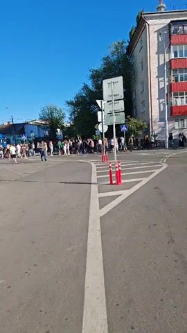 The video shows a street scene with a clear blue sky. A group of people are gathered on the sidewalk and crossing the street. There are road signs, traffic cones, and buildings visible. The people appear to be standing in line or waiting. The perspective suggests the video is taken from a stationary position or moving slowly.