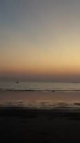 The video shows a sunset at the beach. The sky transitions from a light blue to a yellow-orange hue. The ocean reflects the colors of the sky. There is a small boat in the distance, and a dark silhouette that resembles an island. The beach in the foreground is dark.