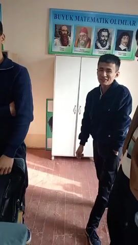 The video shows a young man in a dark blue corduroy jacket, dark pants, and dark shoes, standing in a room with a poster of four mathematicians on the wall. The poster reads "BUYUK MATEMATIK OLIMLAR". There is a white cabinet behind the man and a red-toned wooden floor. He initially smiles at the camera, then puts his hand to his forehead, obscuring his face. He moves his hand down his face, covering it completely. Other people are partially visible on either side of him.
