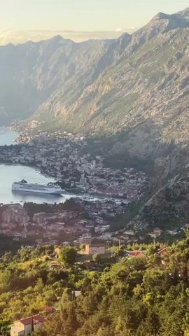 A high-angle shot provides an expansive view of a large bay or fjord, bordered by a town nestled along the coastline and surrounded by majestic, hazy mountains. In the foreground, vibrant green hills are visible, dotted with residential buildings. A large white cruise ship is prominently anchored in the bay. The camera executes a slow, deliberate pan from right to left, revealing more of the extensive waterway, the sprawling town, and a distinctive winding road carved into the steep, vegetated hillside. The sun's soft light illuminates the scene, creating a gentle glow across the water and landscape.