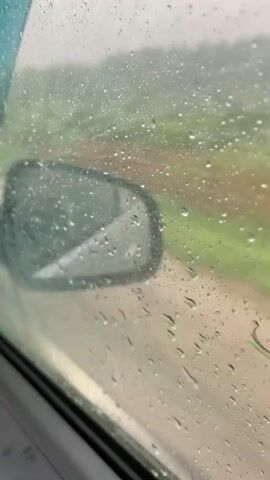 A car window is covered in raindrops, obscuring the view of a road and green landscape outside. A side mirror is visible, reflecting a portion of the car's interior. The camera is positioned inside the car, looking out through the rain-streaked window.